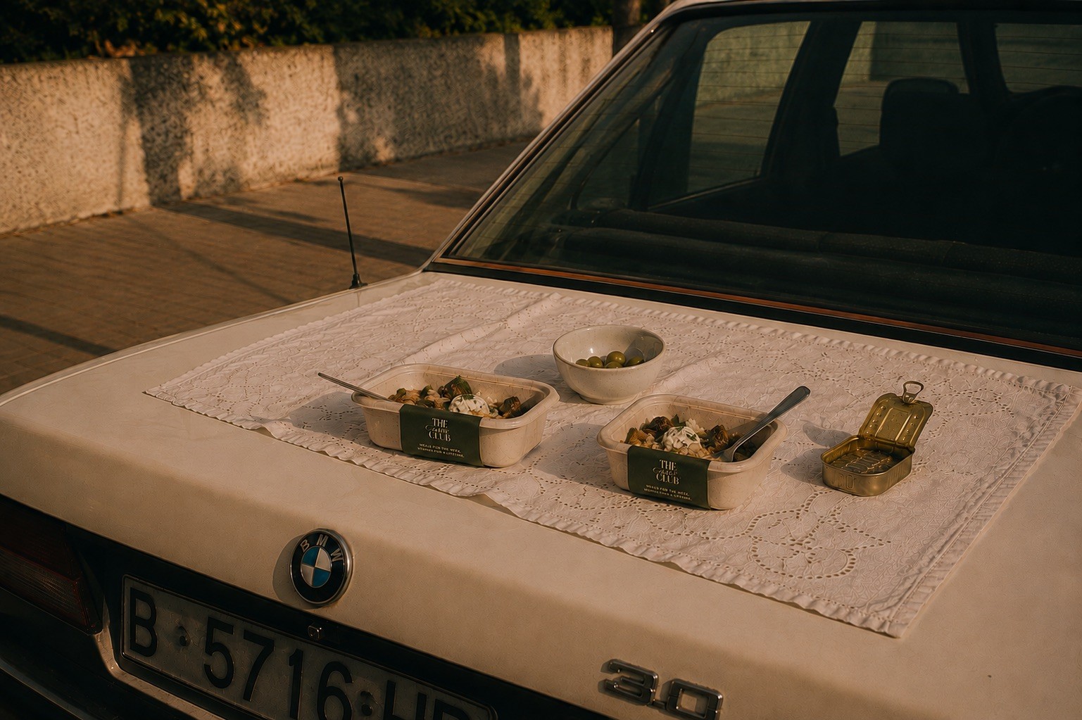 Two Table Club meals, a bowl of olives, and a tin laid on a lace runner across the trunk of a vintage BMW at golden hour